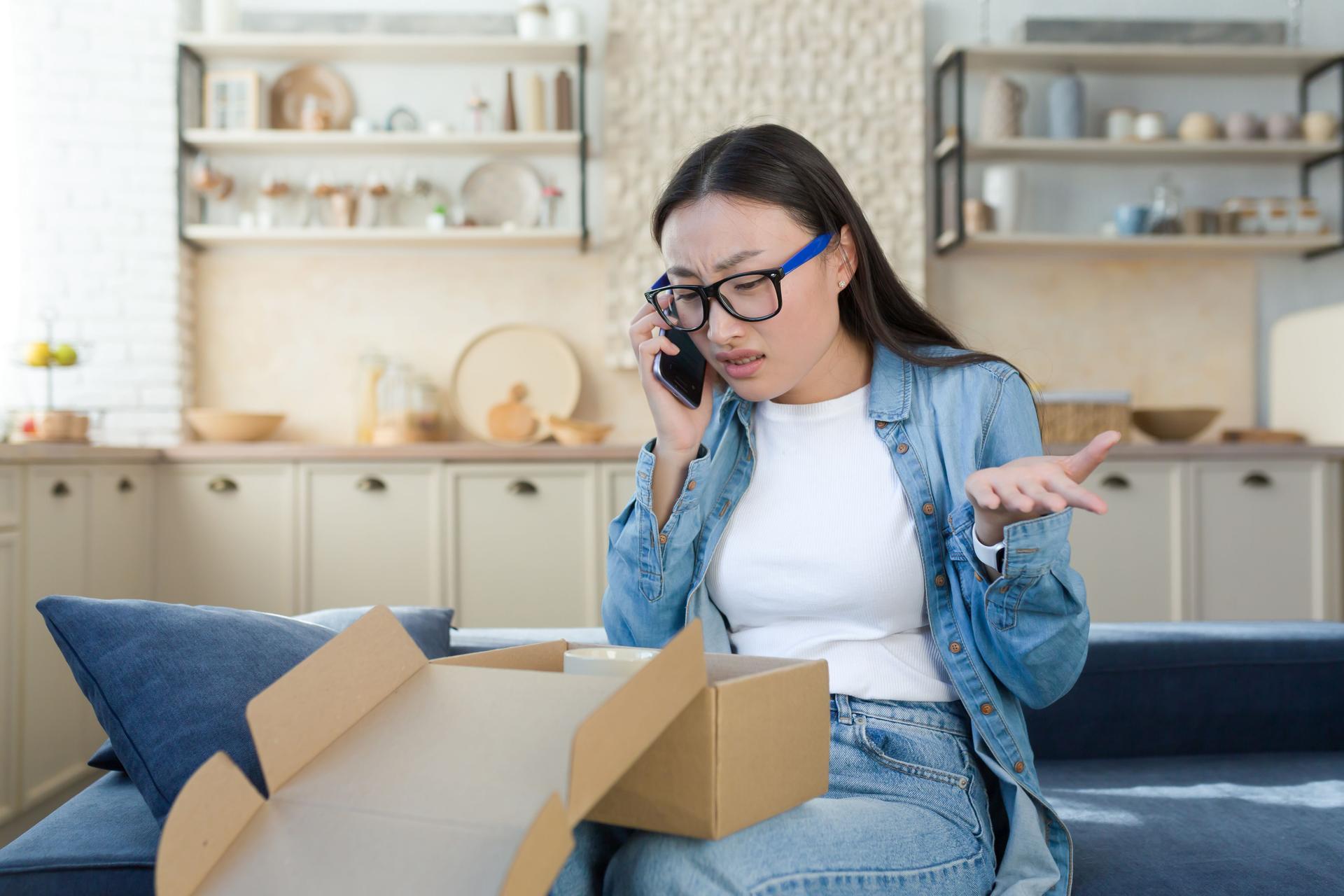 Unsatisfied woman received a broken parcel delivered from an online online store, Asian quarrels with the customer support service, talks to the operator on the phone, sitting home in the kitchen